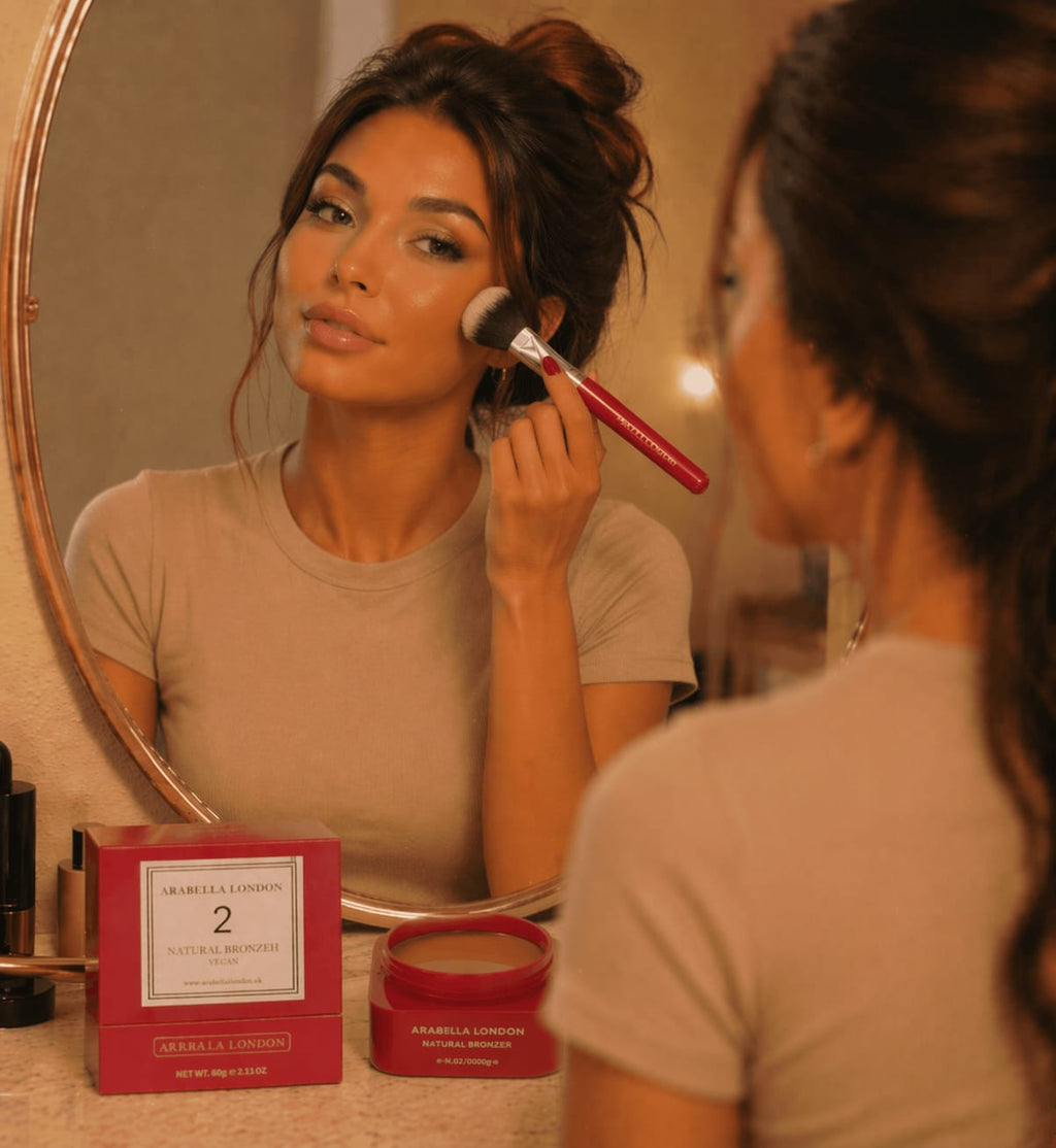 Woman applying Arabella London makeup with a brush in front of a mirror, with a product box and jar on the table.
